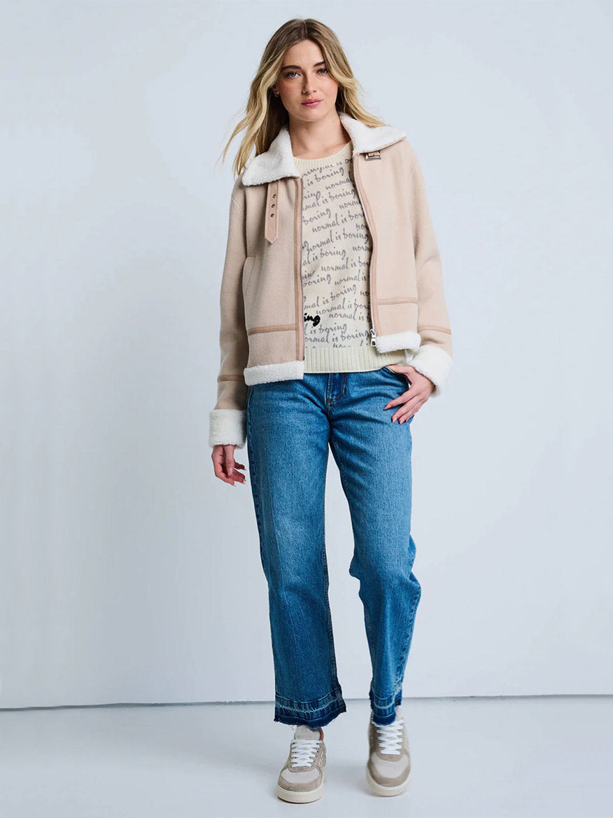 A woman stands against a plain background wearing the Lisa Todd Moto Jacket With Sherpa Trim in Latte/Winter, along with a printed beige top, blue jeans, and beige sneakers.