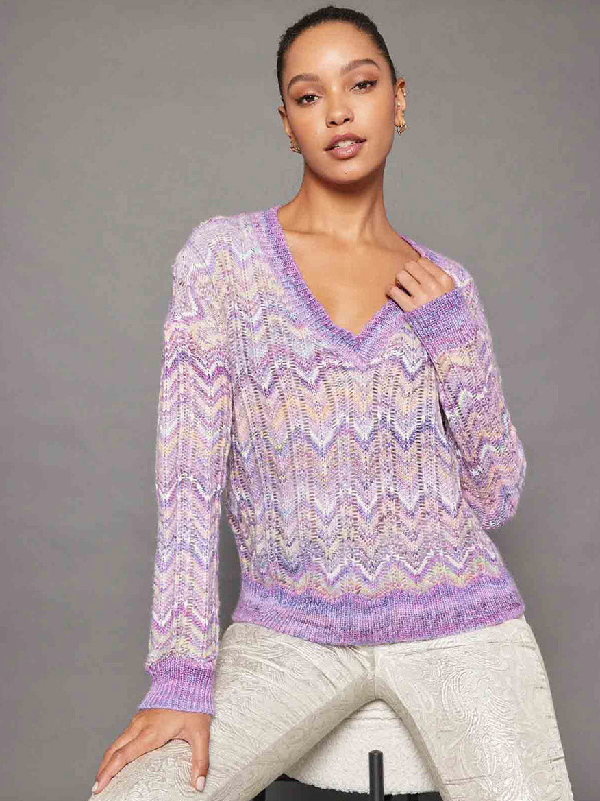 A person wearing the Lisa Todd Mystic Pullover in Purple Haze, which features a multicolored zigzag knit V-neckline, along with light patterned pants, poses against a gray background.