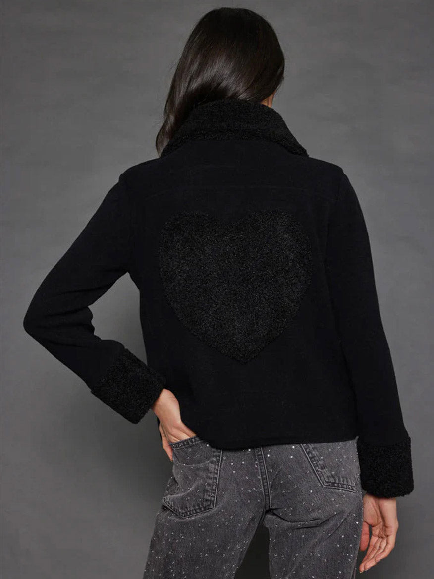 A woman wearing the Lisa Todd No Limits Moto Jacket in black, featuring a textured heart on the back, pairs it with dark jeans while standing against a gray background, facing away from the camera.