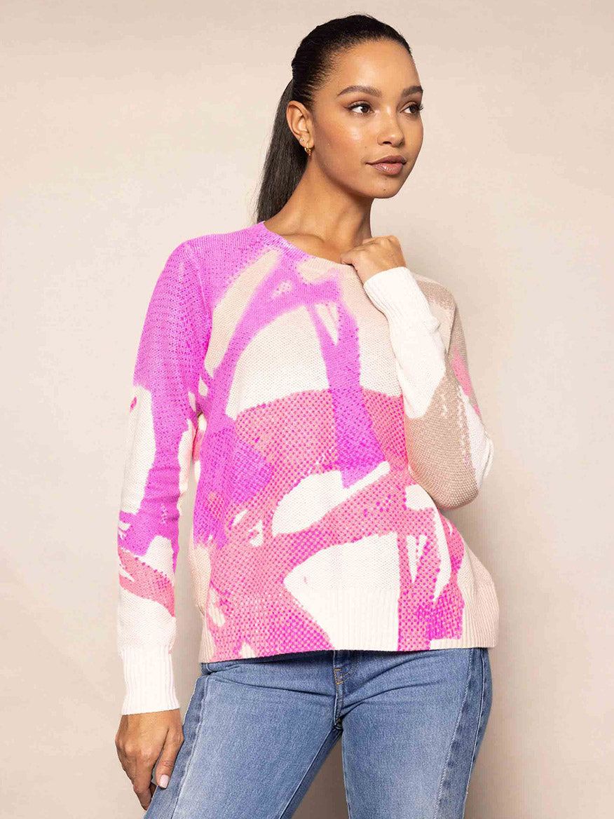 A person wearing the Lisa Todd Painter's Punch Sweater in Pink Pop with blue jeans, standing against a beige background.