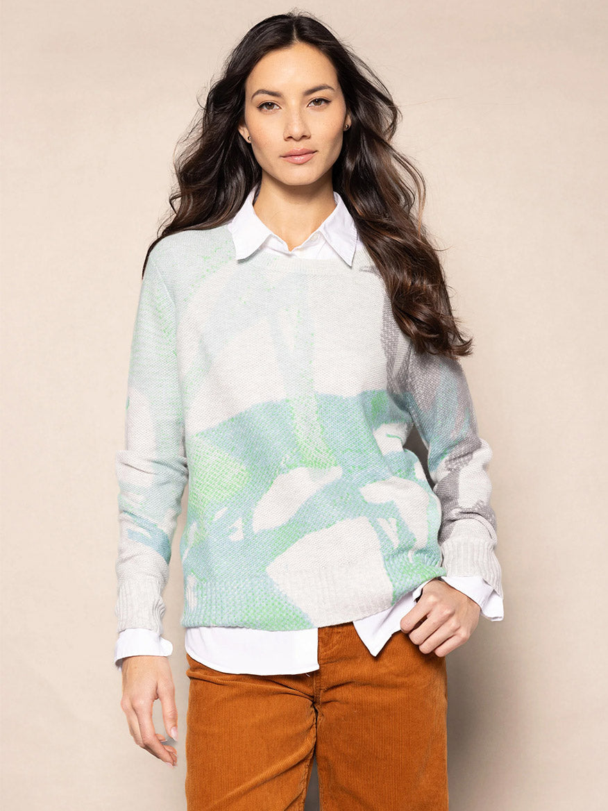 A person wearing the Lisa Todd Painter's Punch Sweater in Blue Hues over a white shirt and brown pants stands against a beige background, evoking the subtle charm of an artistic brush stroke print.