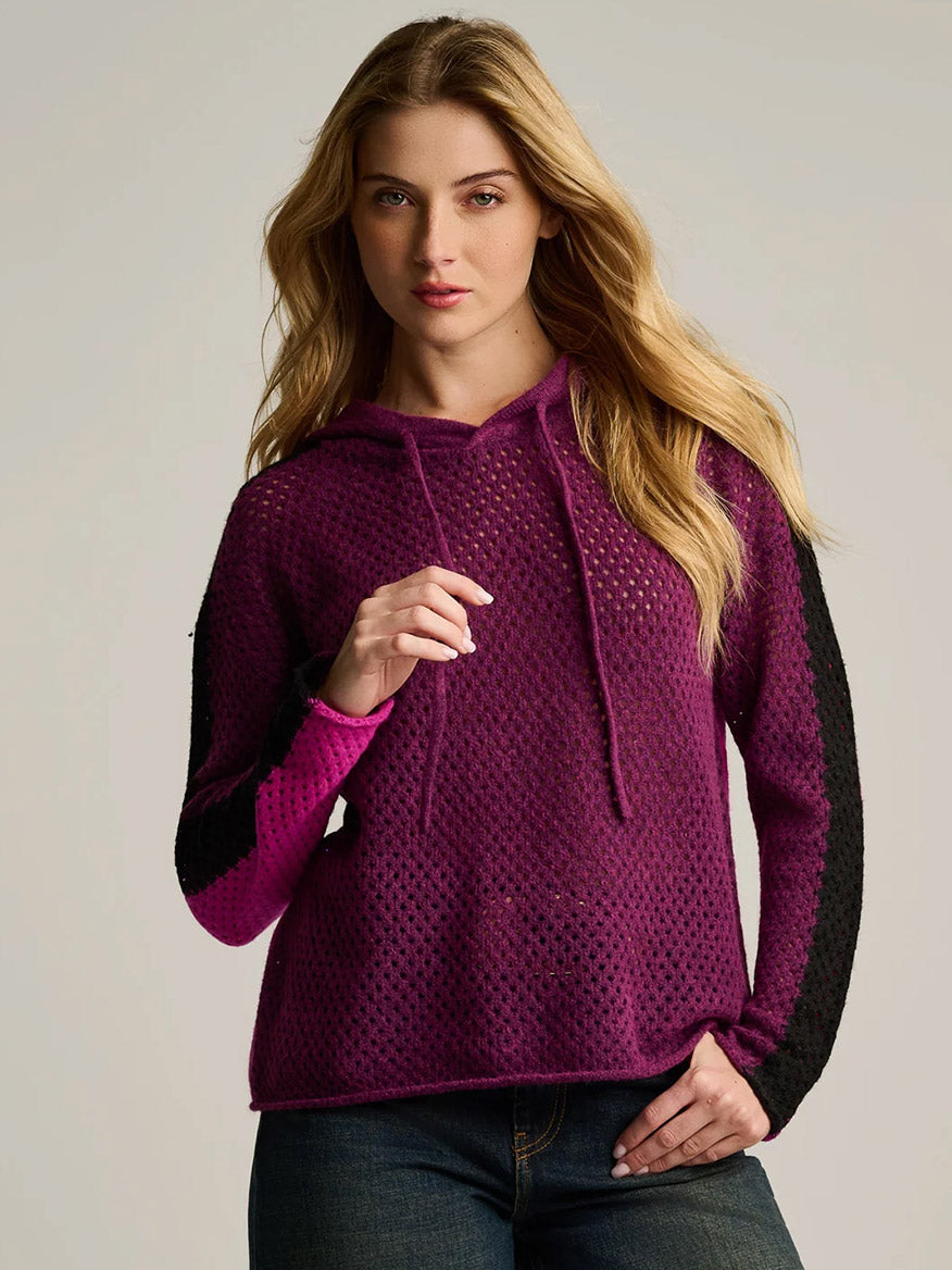 Woman wearing the Lisa Todd Pop Hoodie in Dark Berry Orchid—an oversized hoodie featuring a purple and black palette, perforated texture, and drawstrings, crafted from lightweight knit mesh stitch—standing against a plain light background.