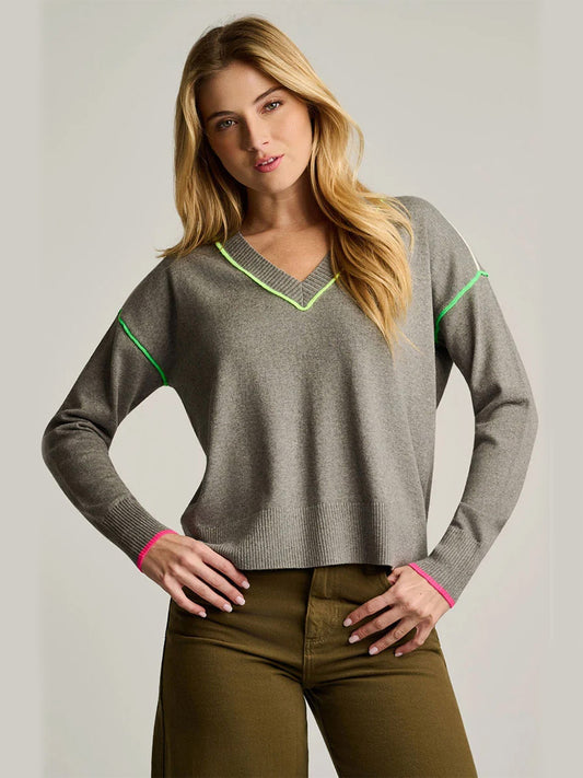 A woman stands against a plain background with one hand on her hip, wearing the Lisa Todd Pop Stix V-Neck Sweater in Grey Shadow—characterized by its relaxed fit, long sleeves, and neon green and pink trim—paired with olive green pants for effortless comfort and style.