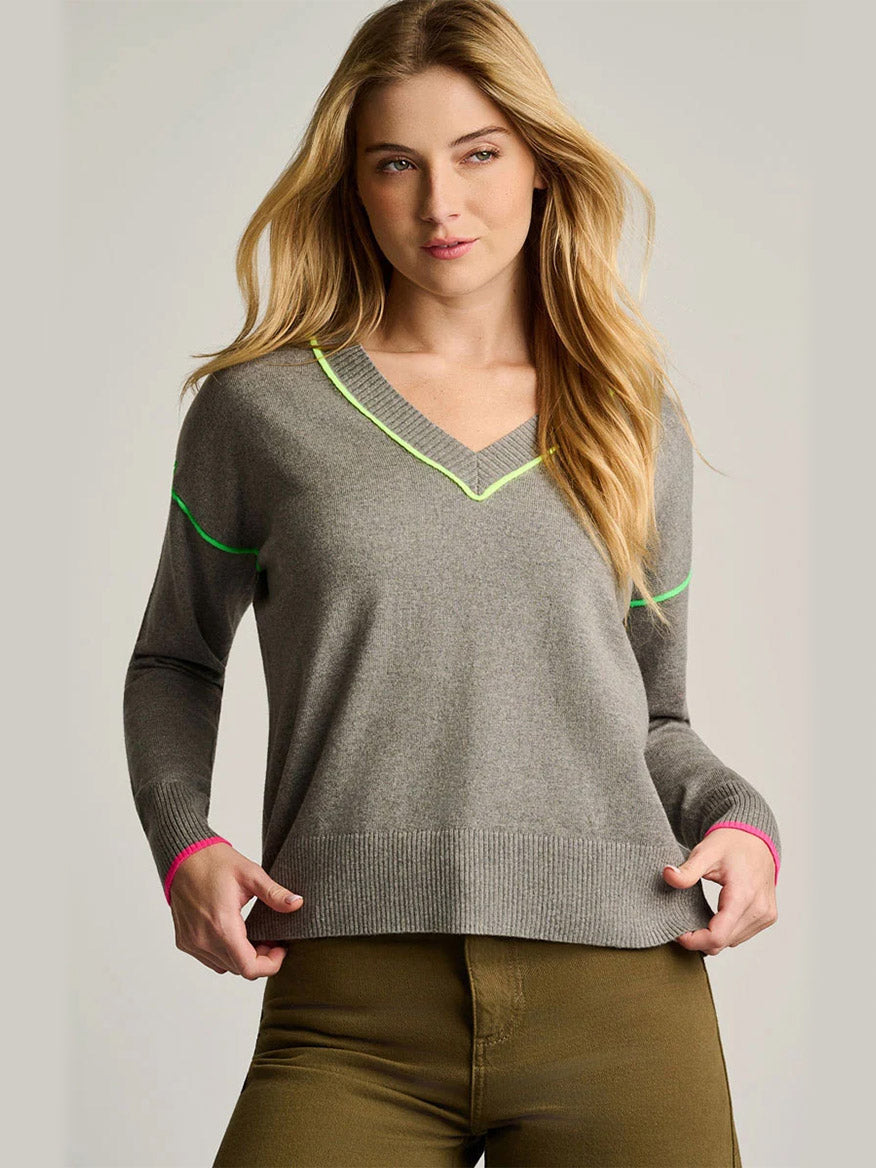 A woman wearing the Lisa Todd Pop Stix V-Neck Sweater in Grey Shadow with neon trim and olive green pants, standing against a plain background.