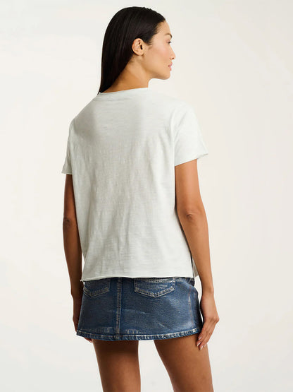 A woman stands facing away, wearing the Lisa Todd Posy Posse T-Shirt in Salty Air and a blue denim mini skirt against a neutral background.