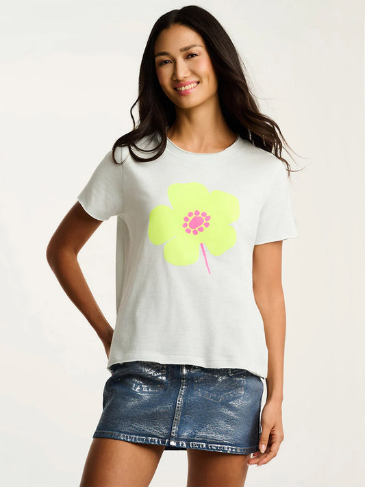 A woman wears the Lisa Todd Posy Posse T-Shirt in Salty Air with a large yellow floral graphic, paired with a metallic blue denim skirt, standing against a plain background.