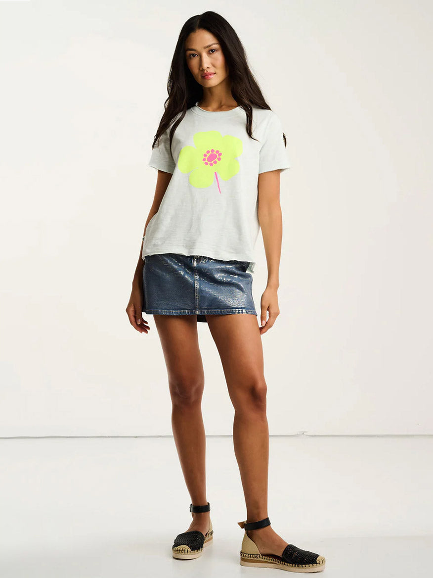 A woman stands in a studio wearing the Lisa Todd Posy Posse T-Shirt in Salty Air, featuring a relaxed fit with a large yellow flower graphic, paired with a metallic blue mini skirt and black espadrille sandals.