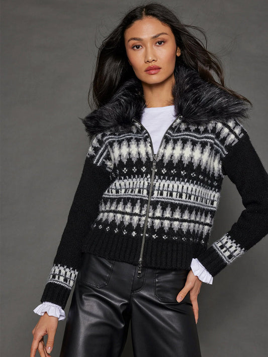 A woman stands against a gray background, elegantly dressed in the Lisa Todd Powder Puff Zip Sweater in Black, featuring a patterned design with a faux fur collar, layered over a white shirt and black leather pants.