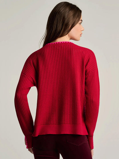 A person wearing a Lisa Todd Power Stitch Cotton Pullover in Spice Red, a relaxed-fit, ribbed red cotton pullover with contrasting pink stitching along the neckline and cuffs.