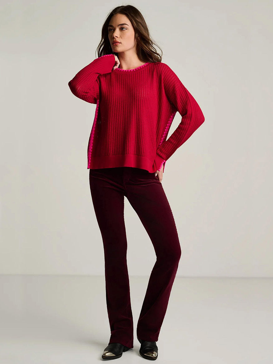 A woman wears a Lisa Todd Power Stitch Cotton Pullover in Spice Red, featuring a relaxed fit, ribbed-knit texture, and pink trim, paired with dark red velvet pants.