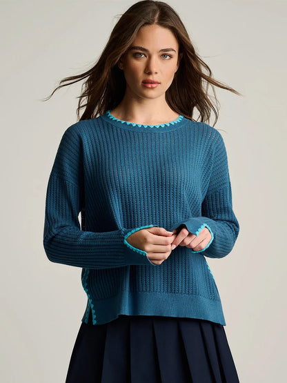 A woman stands against a plain background, facing forward with hands clasped, wearing the Lisa Todd Power Stitch Cotton Pullover in Steel Blue—a ribbed cotton sweater featuring turquoise trim and subtle embroidered accents—paired with a navy skirt.