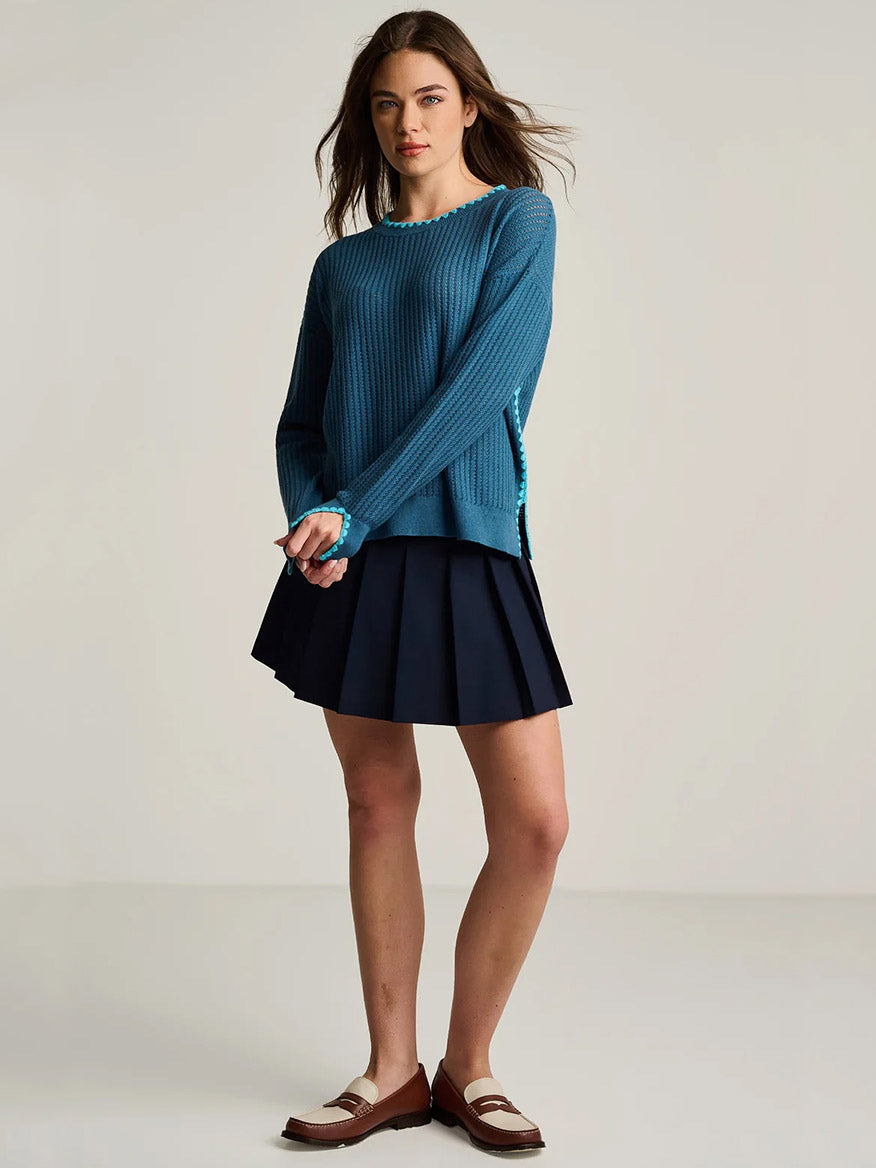 Woman standing in a studio wearing the Lisa Todd Power Stitch Cotton Pullover in Steel Blue, a navy pleated skirt, and brown loafers.