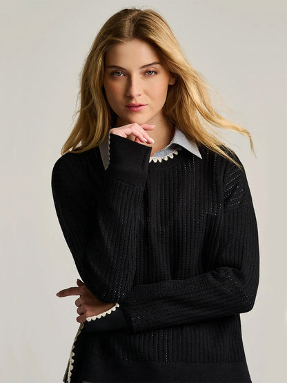 A woman with long blonde hair stands against a plain background, gazing directly at the camera with one hand near her face. She is wearing the Lisa Todd Power Stitch Cotton Pullover in Black, layered over a collared shirt.