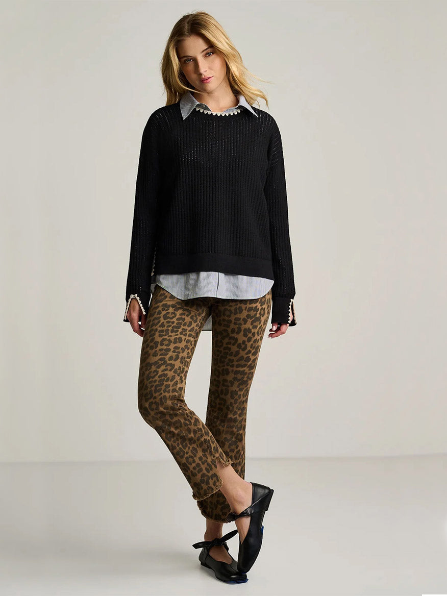 A woman stands against a plain background, wearing the Lisa Todd Power Stitch Cotton Pullover in Black layered over a collared shirt, paired with leopard print pants and black shoes; her legs are crossed and hands by her sides, creating a relaxed yet stylish look.