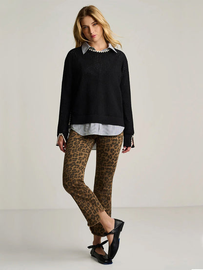 A woman stands against a plain background, wearing the Lisa Todd Power Stitch Cotton Pullover in Black layered over a collared shirt, paired with leopard print pants and black shoes; her legs are crossed and hands by her sides, creating a relaxed yet stylish look.