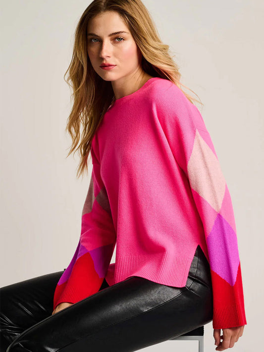 A woman with long blond hair wears the Lisa Todd Power Surge Sweater in Neon Pink featuring bold diamond sleeves and black leather pants, seated against a plain background.