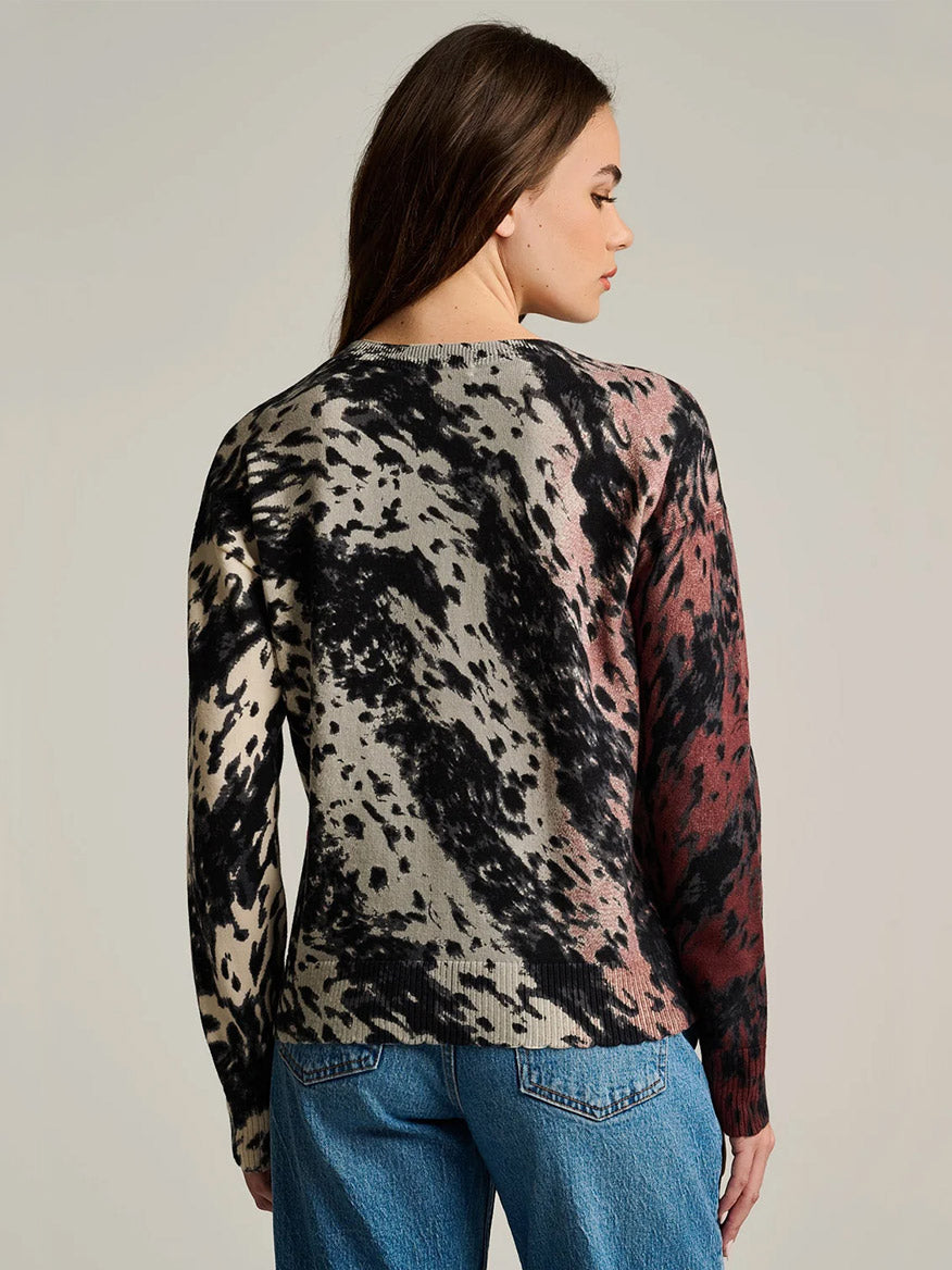 A person with long brown hair, seen from the back, wears the Lisa Todd Running Wild Cotton Sweater in Chocolate Feather—a relaxed fit sweater featuring black, beige, and maroon splatter patterns—paired with blue jeans against a plain background.