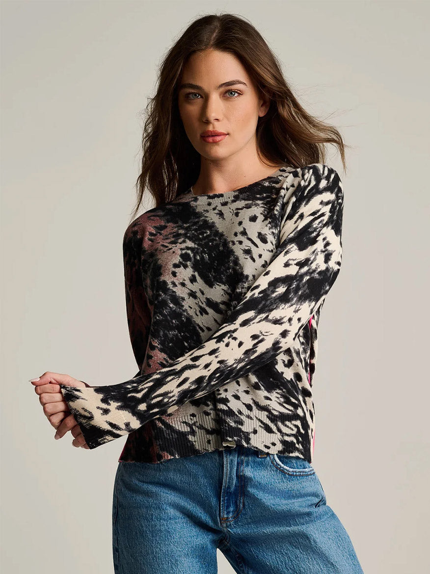 A woman is wearing the Lisa Todd Running Wild Cotton Sweater in Chocolate Feather—a relaxed fit, long-sleeve top featuring a black and white abstract print crafted from 100% cotton—paired with blue jeans and standing against a plain, light background.