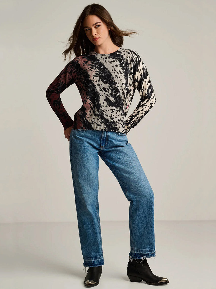 A woman stands in a studio wearing the Lisa Todd Running Wild Cotton Sweater in Chocolate Feather, paired with blue jeans and black ankle boots, with her hands resting on her hips.