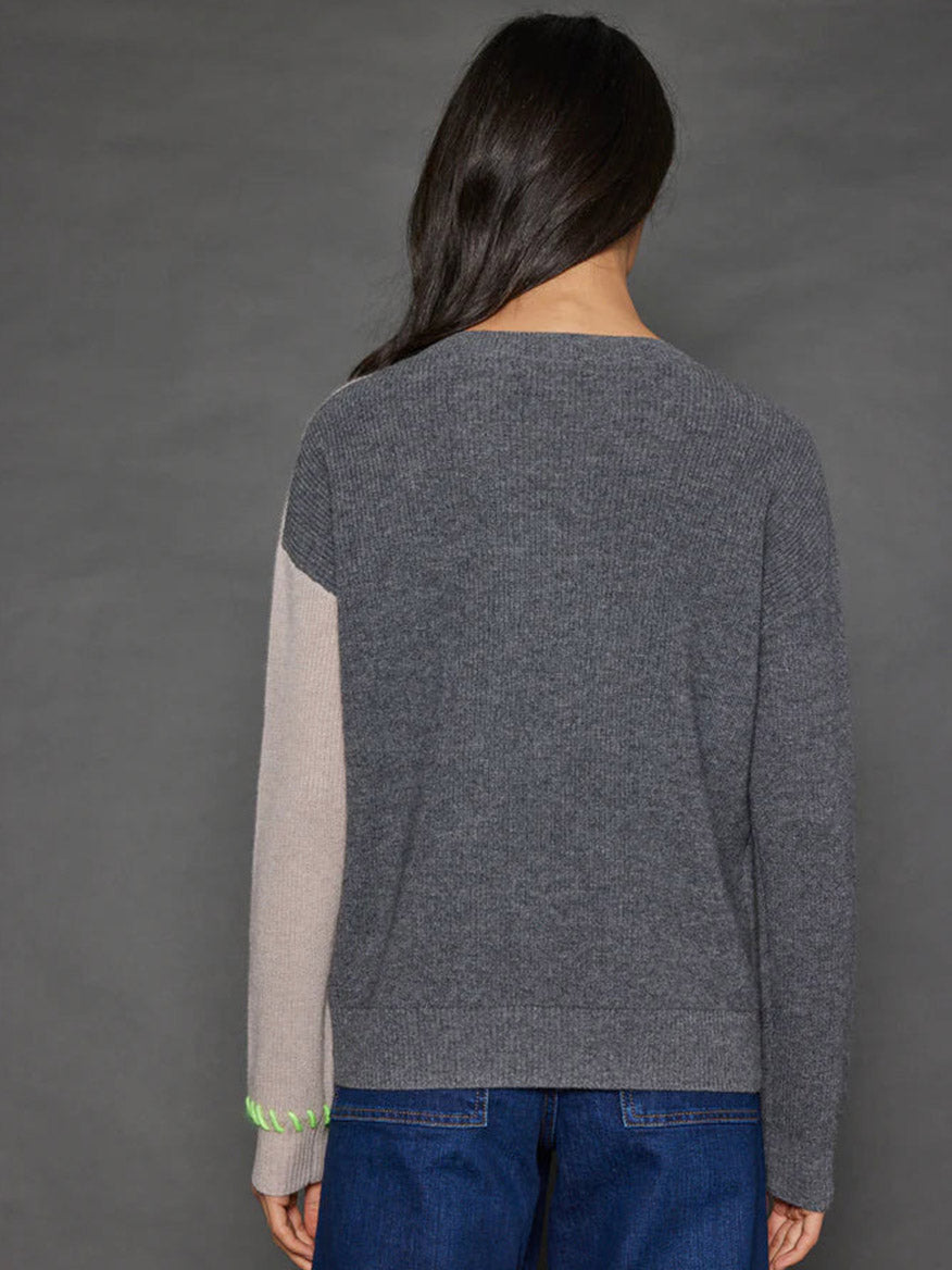 A person with long dark hair is facing away while wearing the Lisa Todd Split Happens Sweater in Chai/Charcoal, a ribbed pullover with an asymmetrical two-tone pattern in gray and beige complemented by neon green stitching, paired with blue jeans.