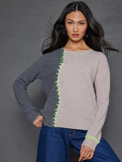 Against a gray background, an individual is dressed in the Lisa Todd Split Happens Sweater in Chai/Charcoal, featuring hand-crafted shaker stitching and neon green accents, seamlessly paired with dark blue jeans.