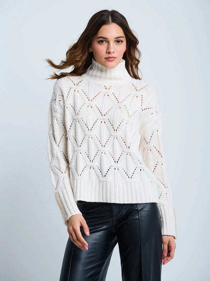 A woman wears the Lisa Todd Stand Out Cable Turtleneck in Winter White with black leather pants, standing against a plain light background.