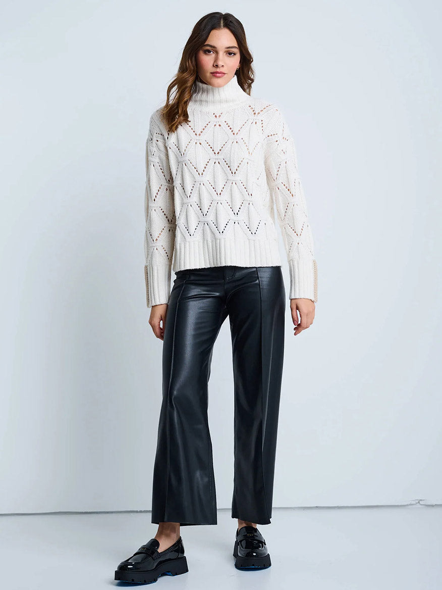 A woman stands against a plain background wearing the Lisa Todd Stand Out Cable Turtleneck in Winter White, styled with black cropped faux leather pants and black platform shoes.