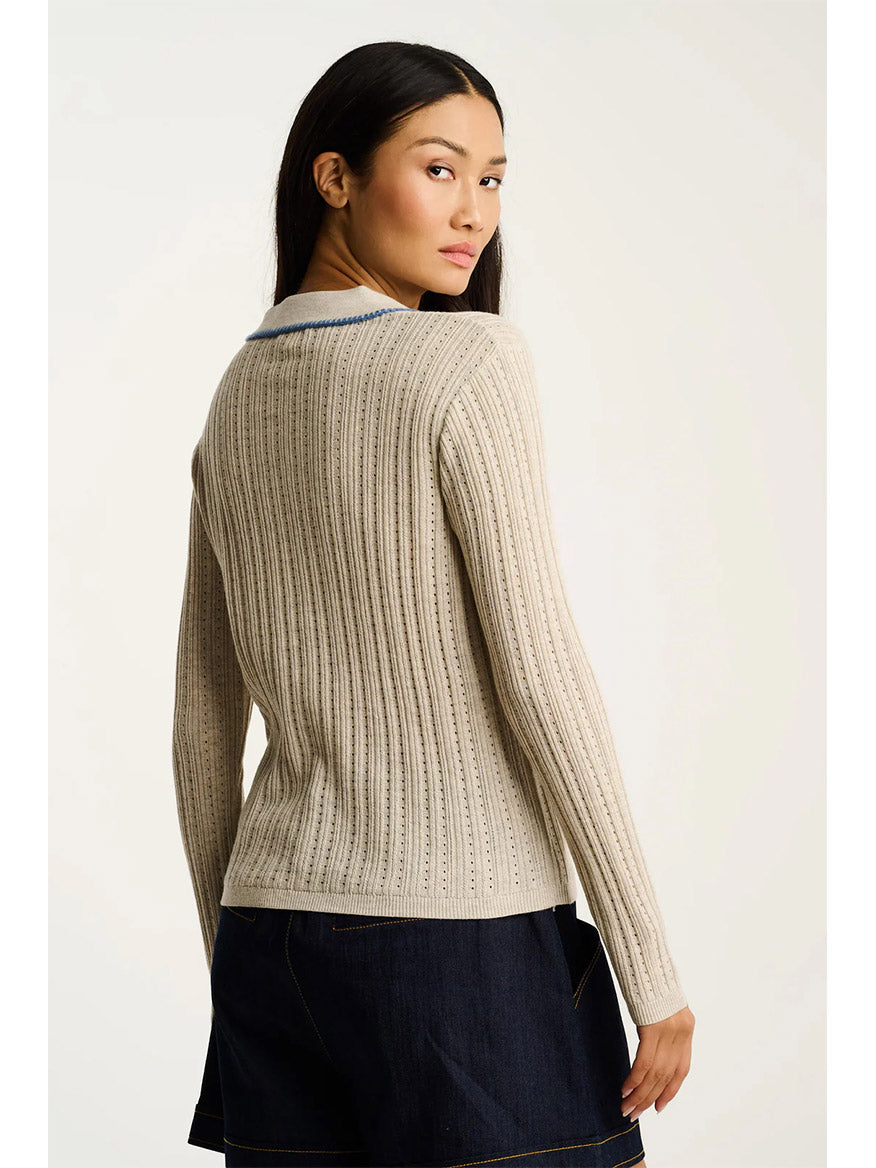 A person with long dark hair wears the Lisa Todd Stitch Please Button-Up Cardigan in Almond, featuring hand-finished rib stitching, paired with a dark skirt, standing with their back to the camera and looking over their shoulder.