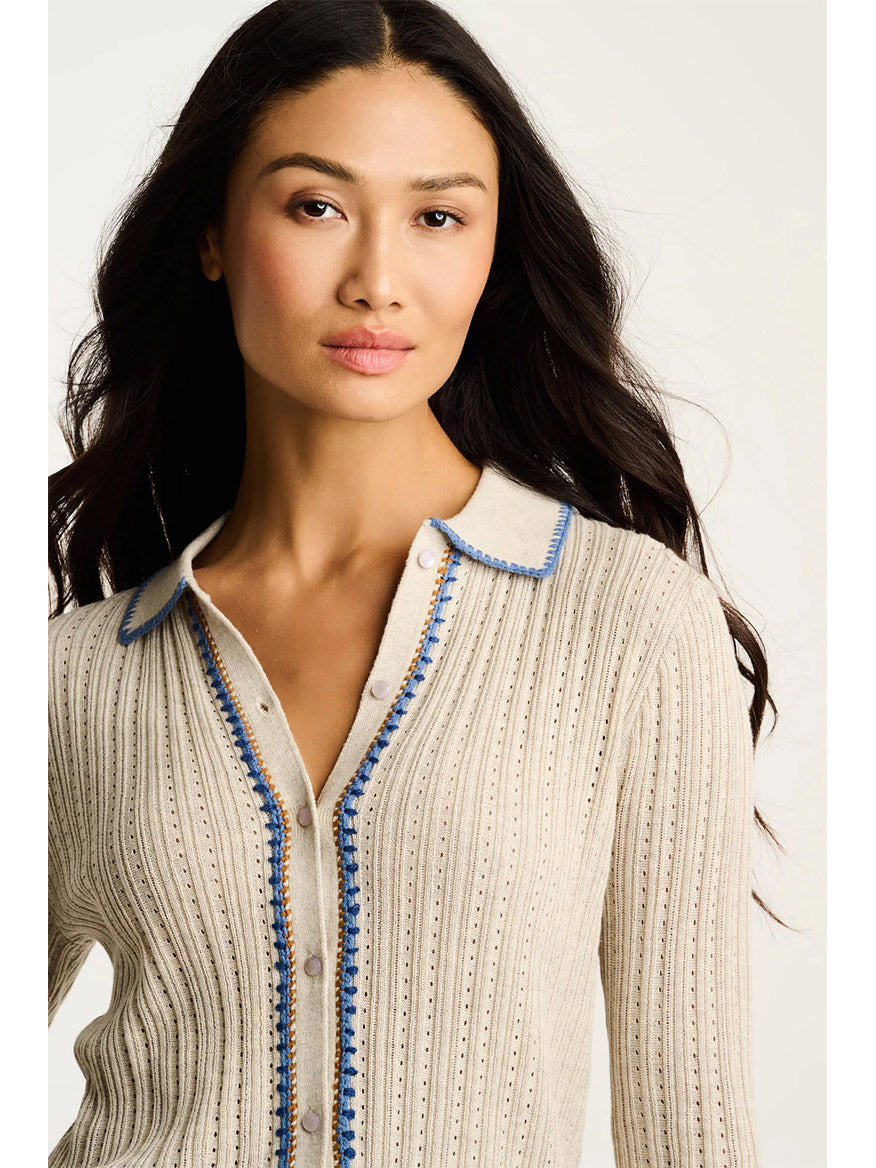 A woman stands against a plain light background, wearing the Lisa Todd Stitch Please Button-Up Cardigan in Almond, featuring blue trim along the collar and button placket with delicate rib stitching.