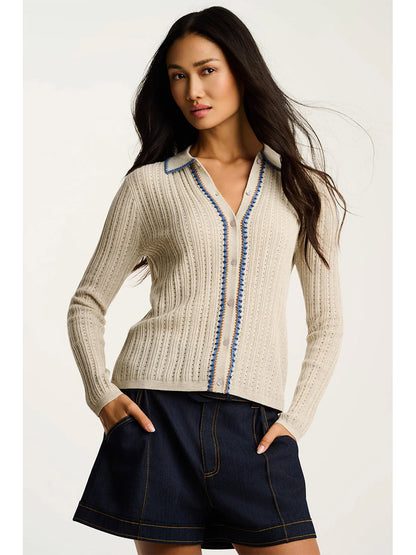 A woman wears the Lisa Todd Stitch Please Button-Up Cardigan in Almond, which features blue hand-finished contrast trim and rib stitching, paired with dark denim shorts as she stands with hands in her pockets against a plain background.