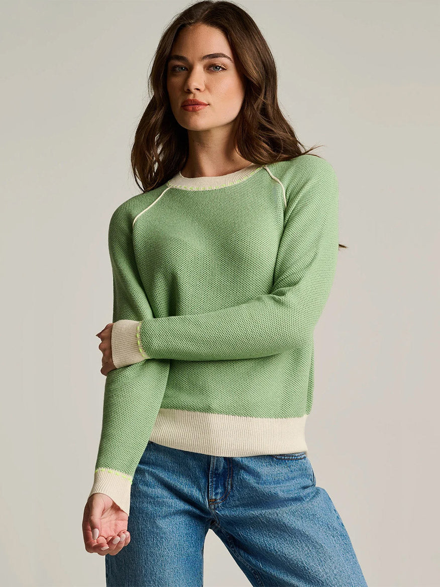 A woman stands with her arms crossed against a neutral background, wearing the Lisa Todd Stitchin’ Cotton Cashmere Sweater in Sage—a light green sweater with cream trim, crafted from cotton-cashmere and featuring an artisanal finish—paired with blue jeans.