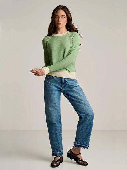 A woman stands against a plain background, wearing the Lisa Todd Stitchin’ Cotton Cashmere Sweater in Sage, paired with blue jeans and black woven shoes.