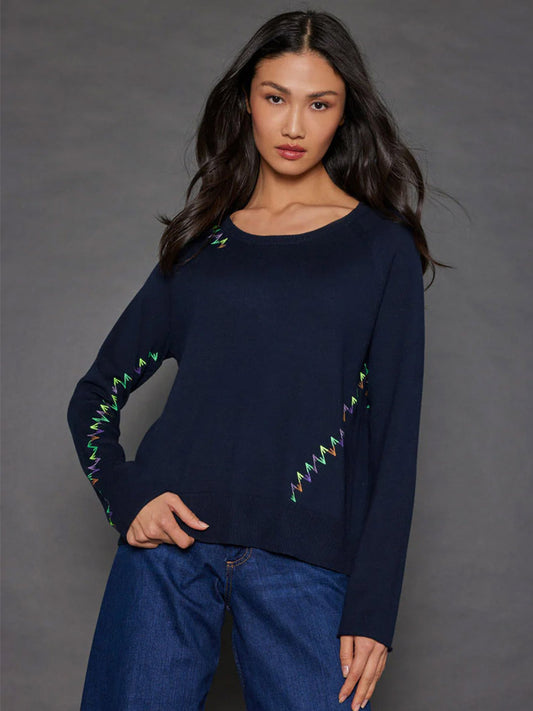 A person stands against a dark background, wearing the Lisa Todd Stitchmix Sweater in Ink, featuring colorful zigzag patterns on the sleeves and shoulders, paired with blue jeans.