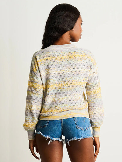 A woman faces away, wearing the Lisa Todd Sun-Sational V-Neck Pullover in Watercolor with yellow accents and space-dye pattern, paired with frayed denim shorts against a plain background.