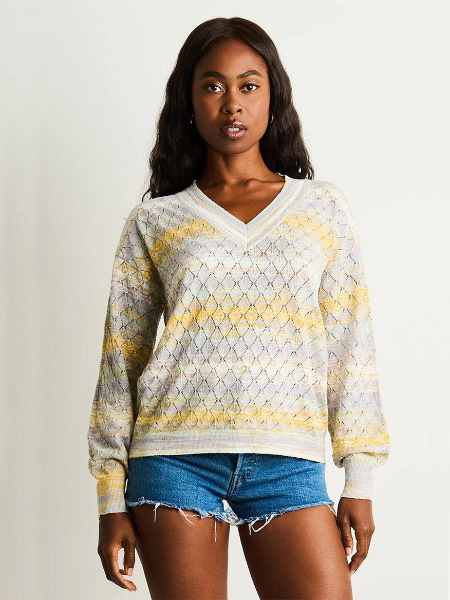 A woman faces forward, wearing the Lisa Todd Sun-Sational V-Neck Pullover in Watercolor with a yellow and gray space-dye pattern and blue denim shorts against a plain light background.