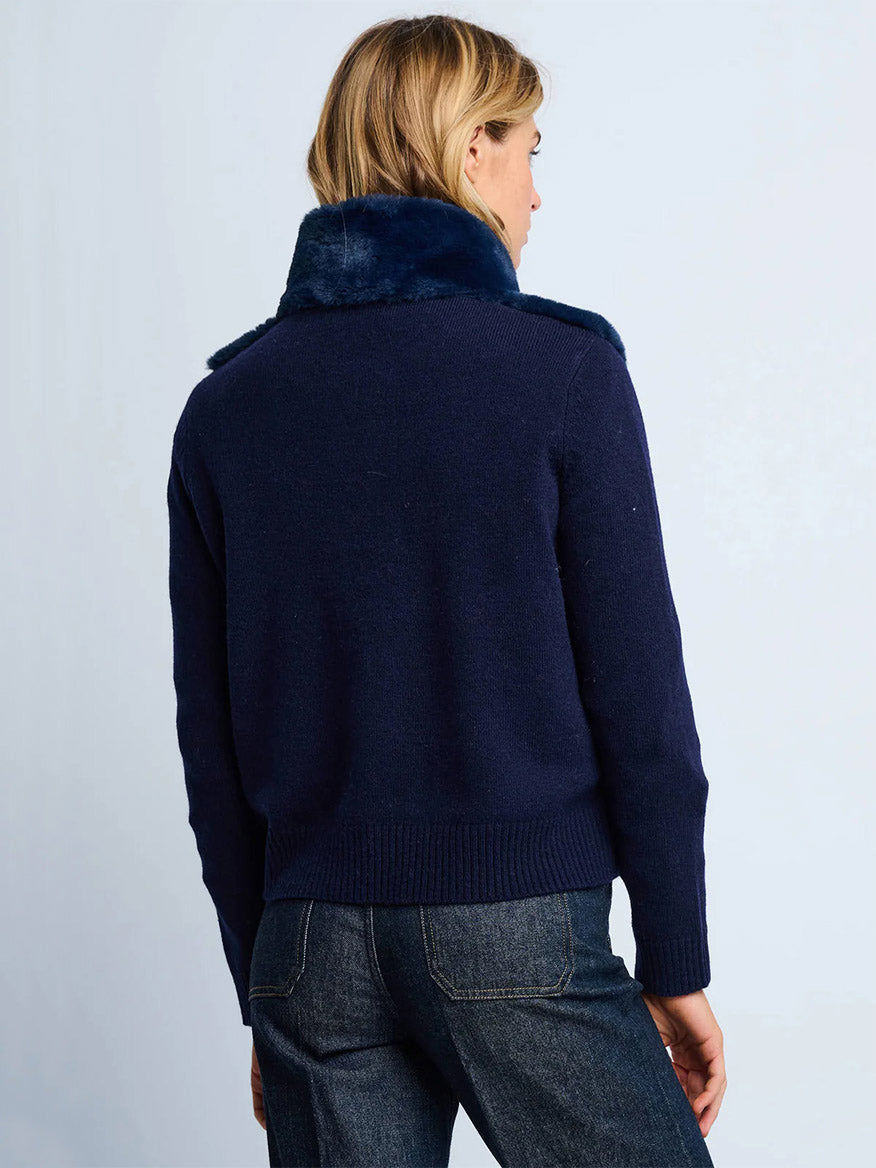 A person with blonde hair is seen from the back, wearing the Lisa Todd Teddy Faux Fur Knit Jacket in Navy with a plush collar, paired with dark denim jeans.