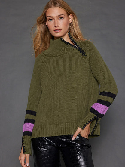 A person wearing the Lisa Todd Unzipped Sweater in Eden with chunky knit and black and purple accents, paired with black textured pants, stands against a gray background, highlighting the contrast embroidery detail.