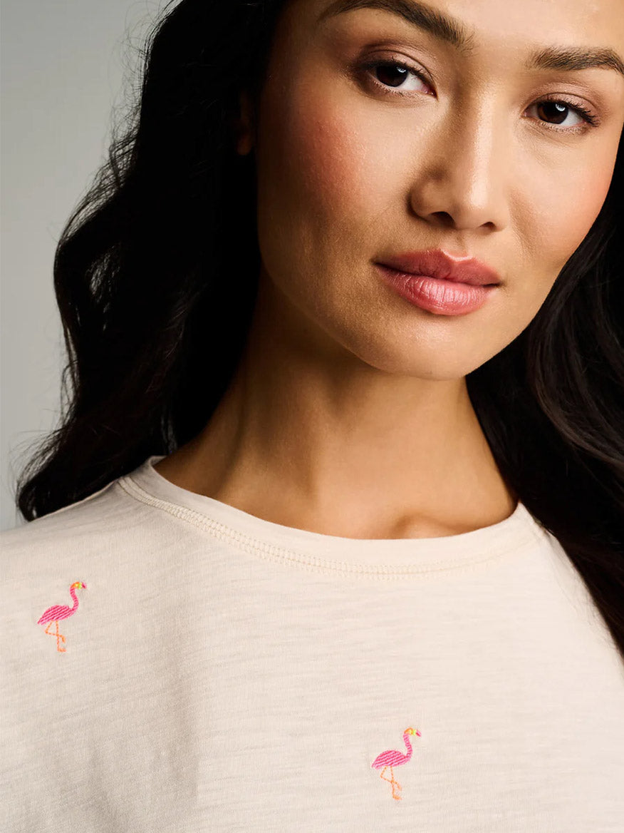 A woman wears the Lisa Todd What The Flock Tee in Crystal Sand, a relaxed cotton top featuring two small pink flamingos embroidered on the front.