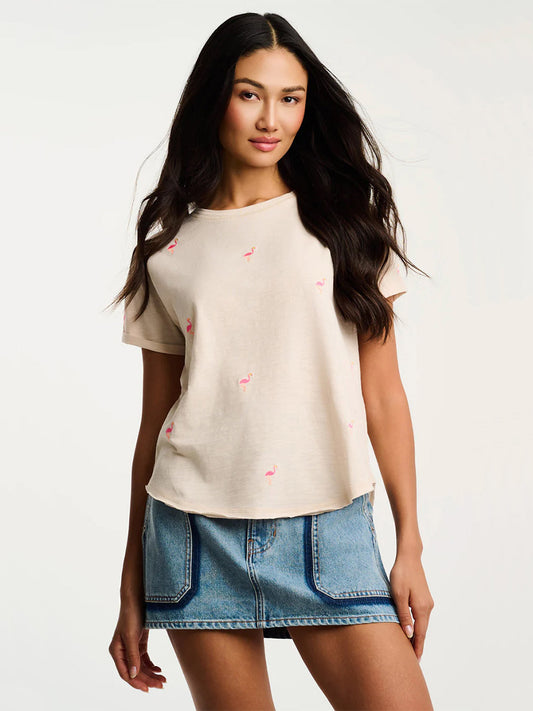 Woman wears the Lisa Todd What The Flock Tee in Crystal Sand, a relaxed cotton top with flamingo embroidery, paired with a denim mini skirt against a plain white background.