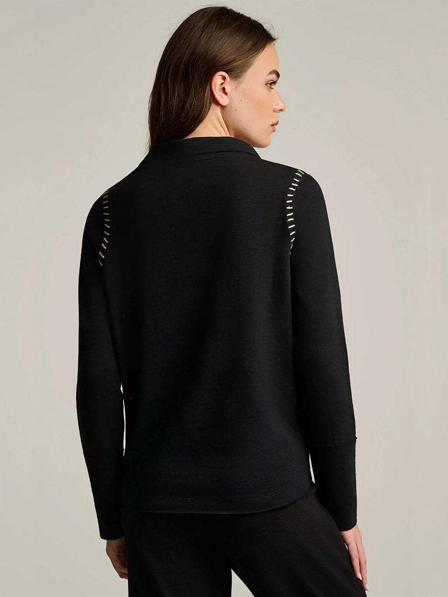A woman stands with her back to the camera, wearing the Lisa Todd Whipster Cotton T-Shirt in black. The relaxed-fit top features white stitch details and whipstitch trim on the shoulders, paired with black pants.