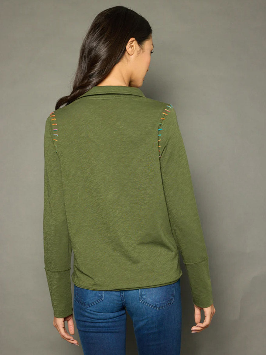 A woman with long brown hair is standing facing away, wearing the Lisa Todd Whipster Long Sleeve Polo in Jungle with multicolored lace details on the shoulders and blue jeans featuring contrast whip stitch details.