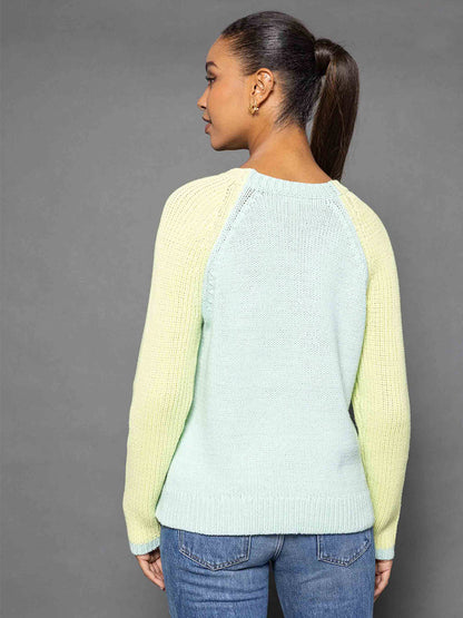 A woman with a ponytail is wearing the Lisa Todd Zip Me Sweater in Ice Combo, which features a light green and yellow color block design, paired with blue jeans as she stands sideways against a gray background.