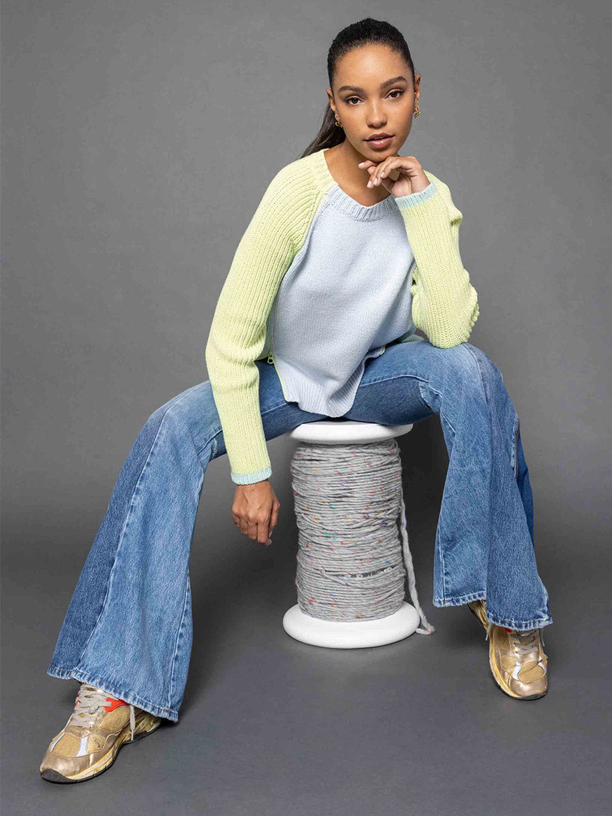 A person wearing the Lisa Todd Zip Me Sweater in Ice Combo, blue jeans, and sneakers sits on a spool of thread against a gray background.