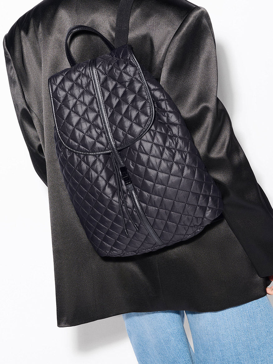 A person is photographed from behind wearing the MZ Wallace Astor Backpack in Black Oxford, featuring gunmetal hardware, over a black blazer and light blue jeans, set against a plain white background.