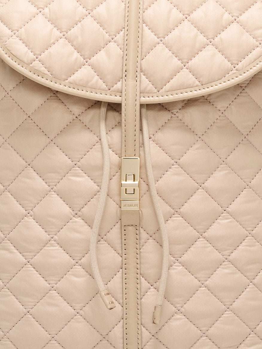 Close-up of the MZ Wallace Astor Backpack in Buff Oxford, featuring beige quilted leather, a drawstring closure, and a gold-tone central buckle that highlights its luxe detailing.