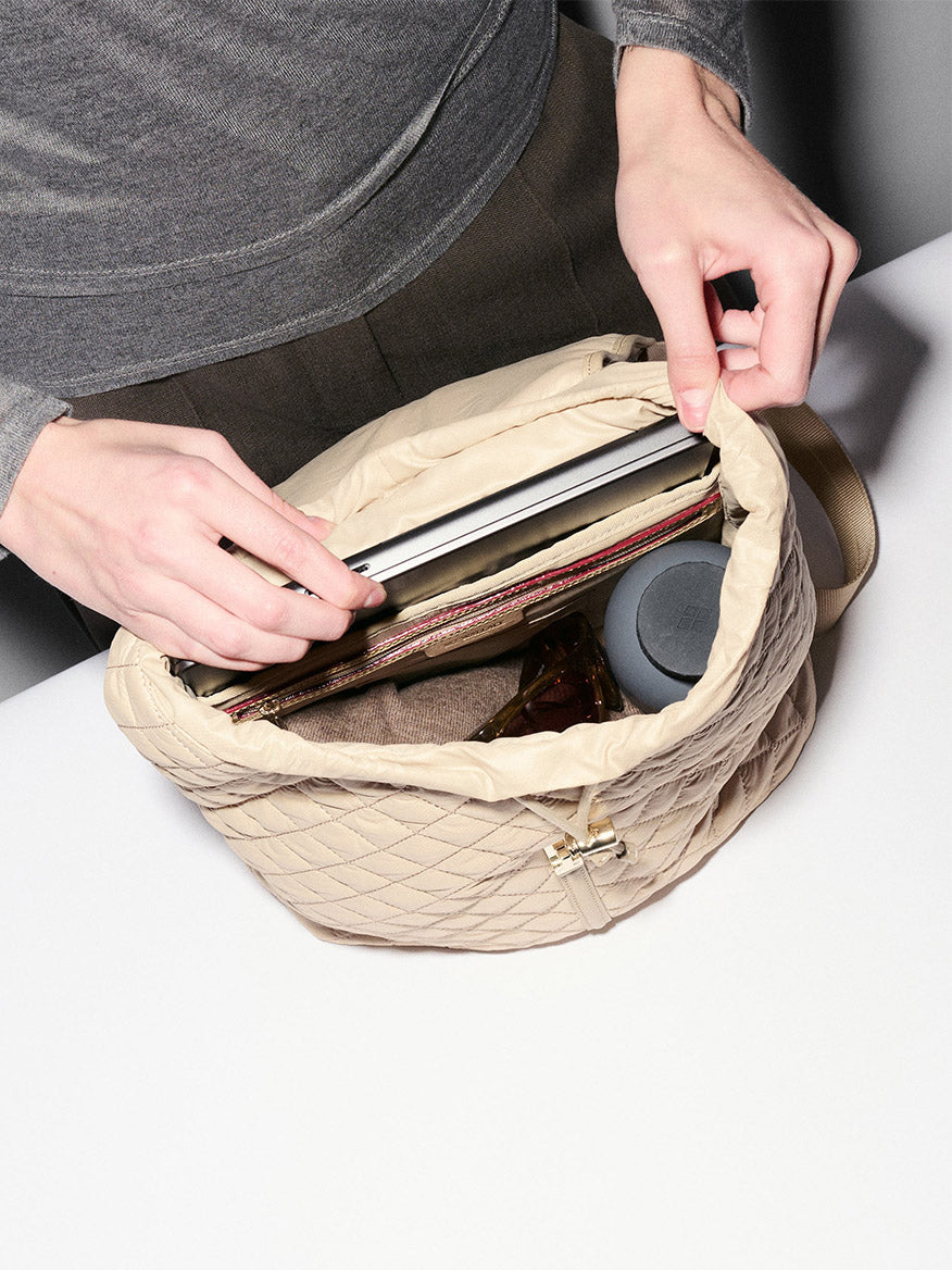 A person places a laptop into an MZ Wallace Astor Backpack in Buff Oxford, which also holds a small round case and a dark bottle.