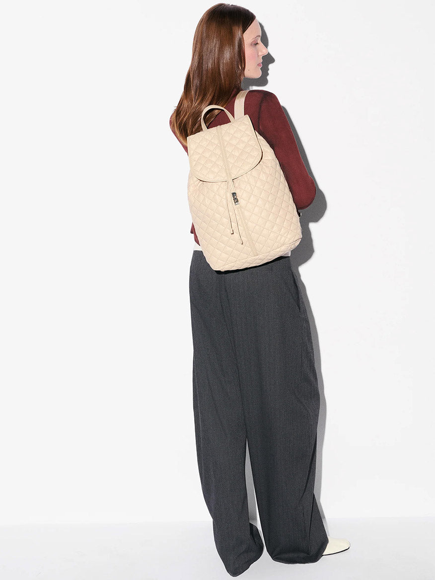 A woman stands sideways, wearing a maroon long-sleeve top, gray wide-leg trousers, and carrying the MZ Wallace Astor Backpack in Buff Oxford—a beige quilted laptop backpack.