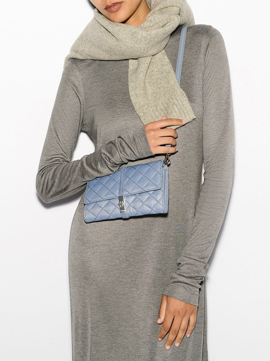 A person wearing a grey dress and beige scarf, accessorized with the MZ Wallace Astor Convertible Crossbody in Stone Blue Oxford featuring an elegant Italian leather strap.