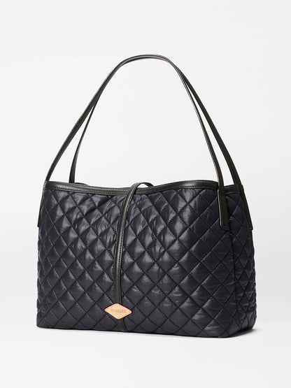 The MZ Wallace Astor East West Tote in Black Oxford features black quilted Italian leather, double shoulder straps, gunmetal hardware, and a gold emblem on the front, shown against a plain white background.