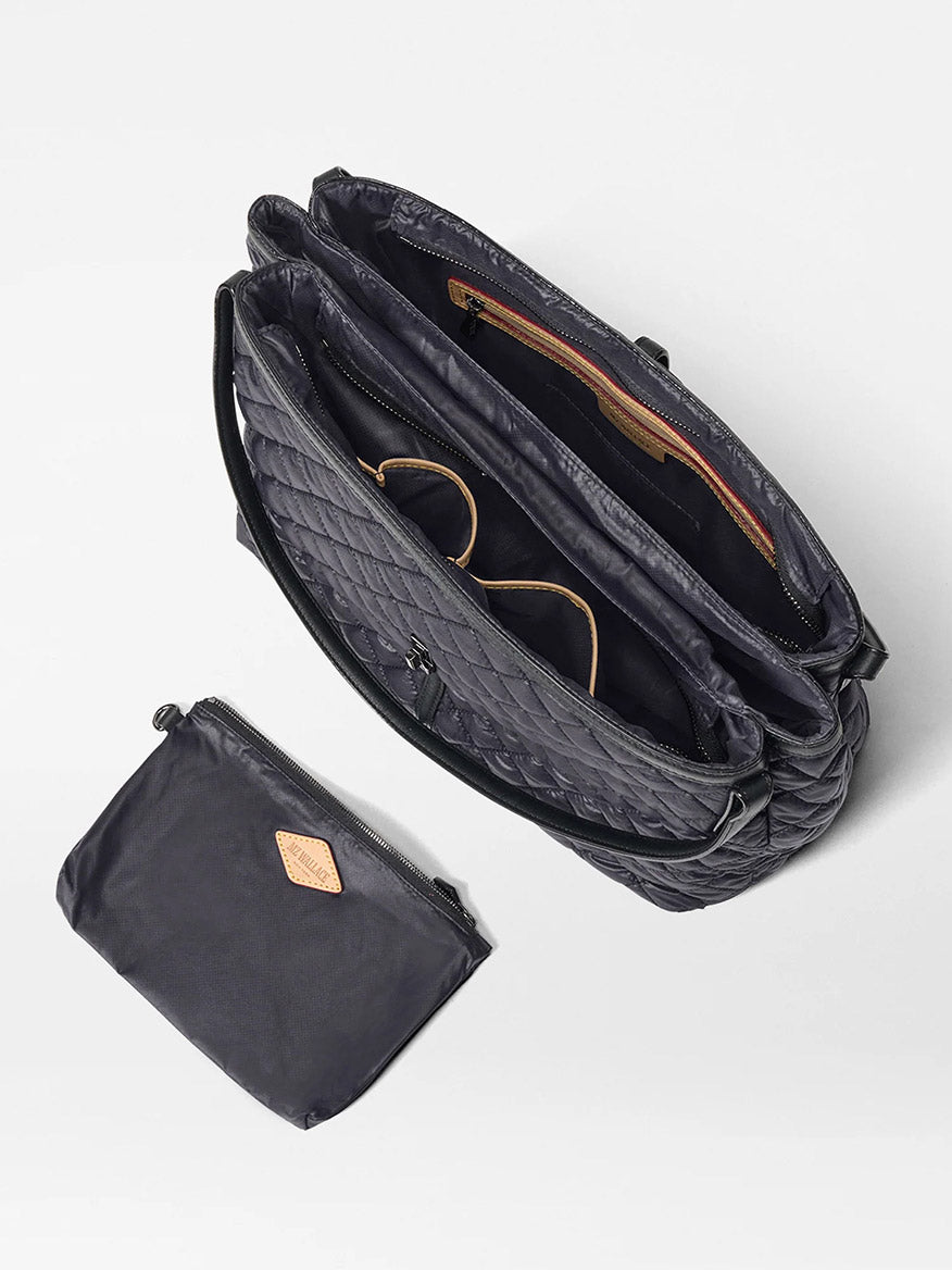The MZ Wallace Astor East West Tote in Black Oxford, featuring gunmetal hardware and multiple compartments, is displayed from above beside a matching black zippered pouch on a white background.